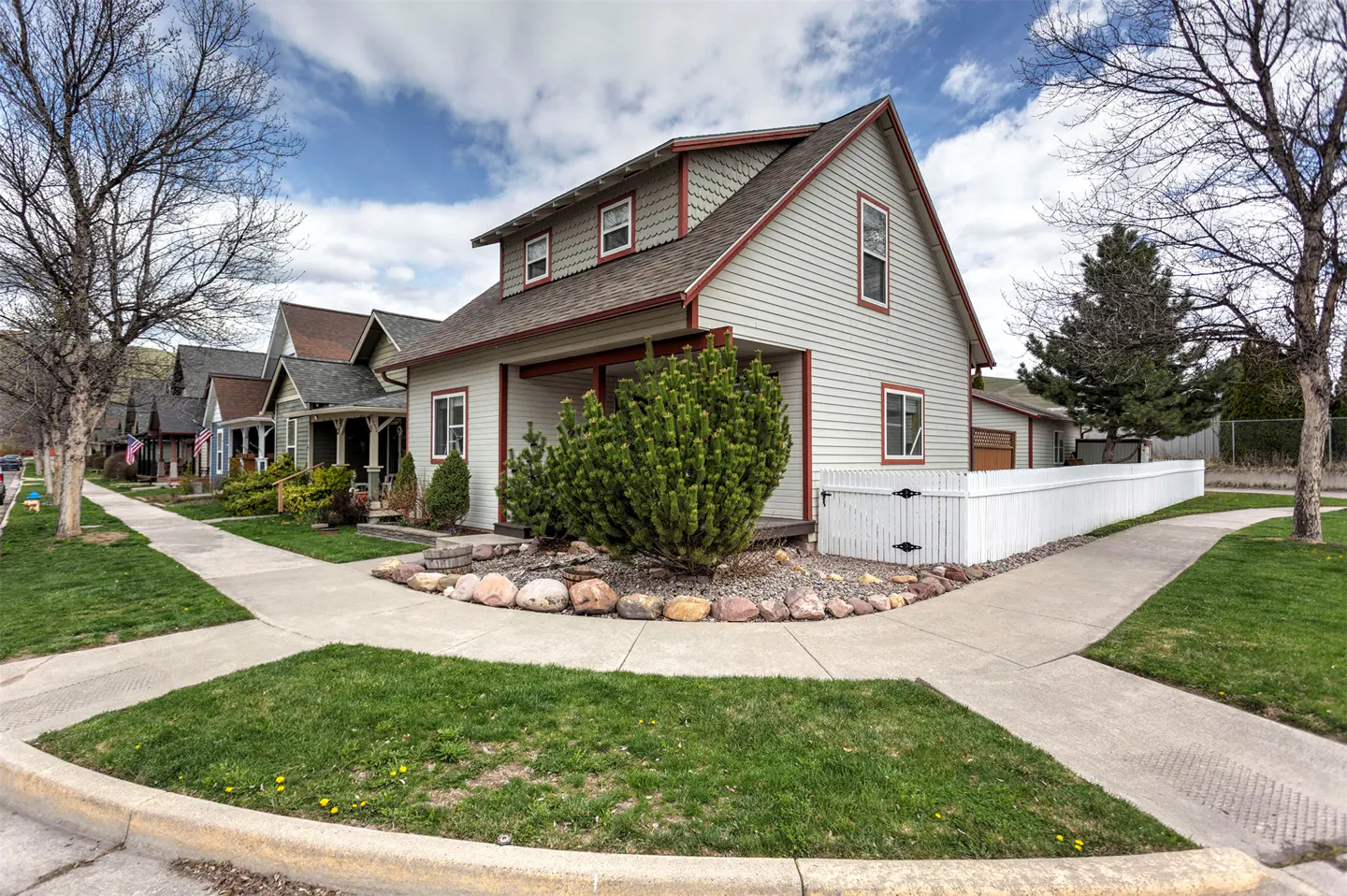 A light gray two-story house with a red trim and a brown roof, surrounded by green bushes and a white fence.