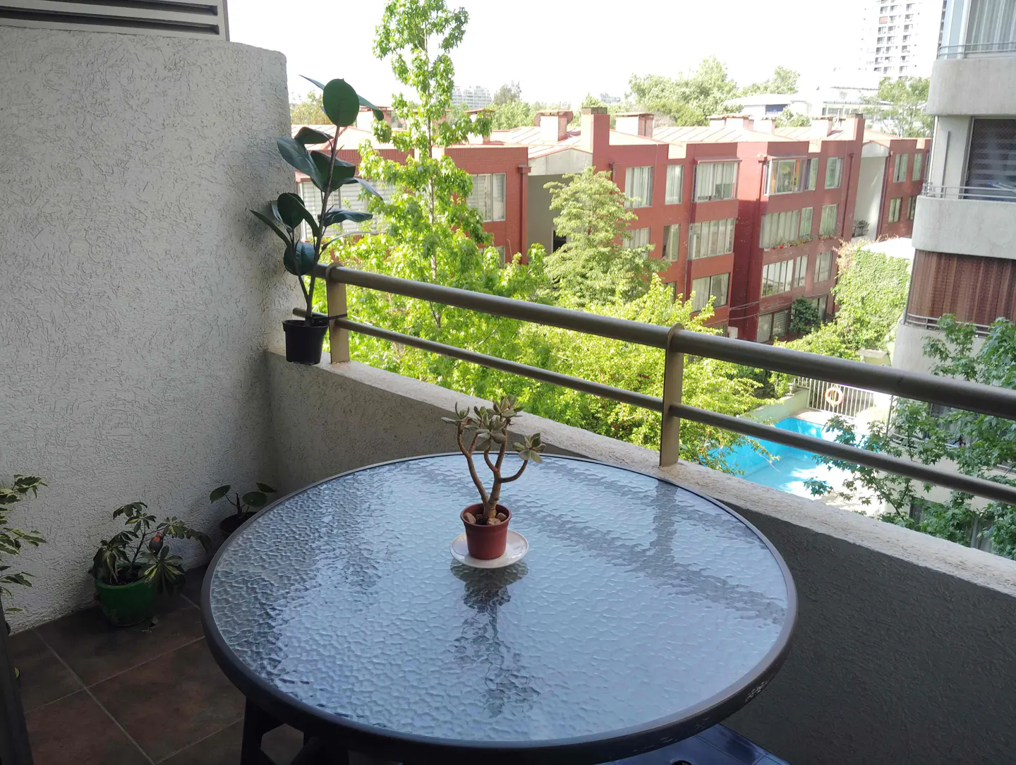 Balcony view with a glass table, potted plants, and a view of red brick buildings and a blue swimming pool.