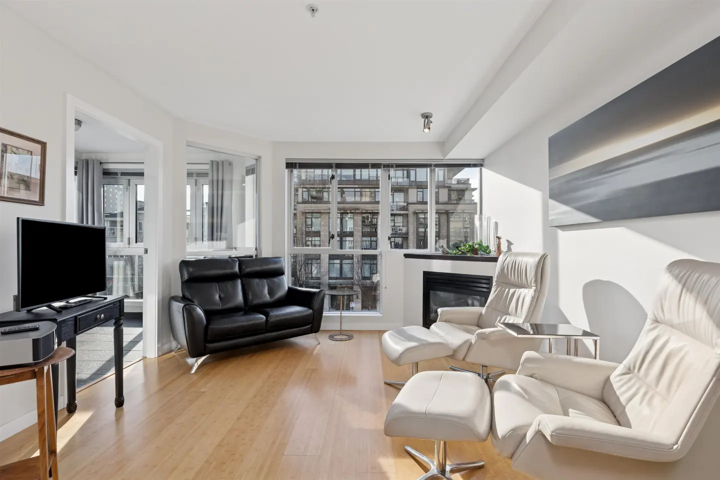 Bright living room with wood floors, black leather sofa, and two white leather recliners with ottomans. A fireplace and city view are visible.