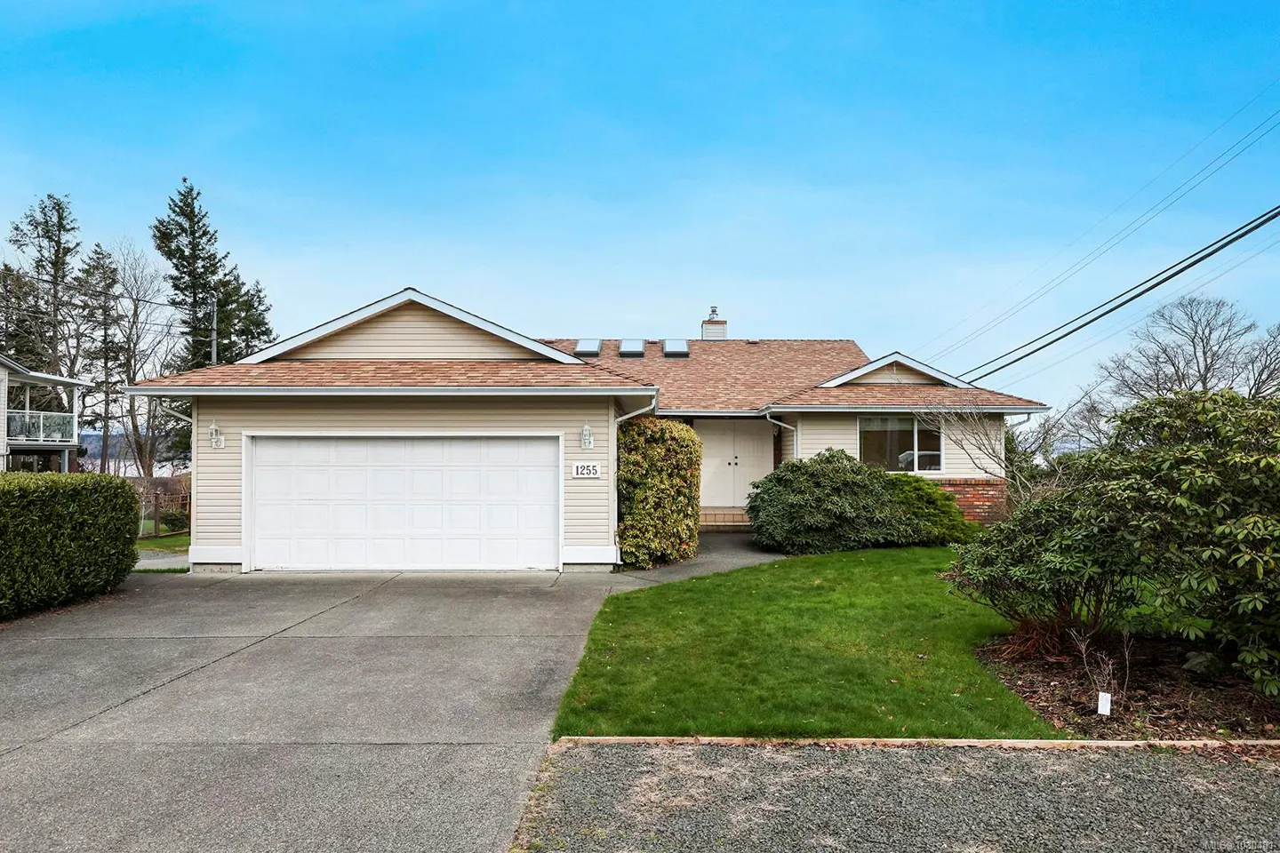 A single-story beige house with a brown roof, a white garage door, and green bushes under a blue sky.