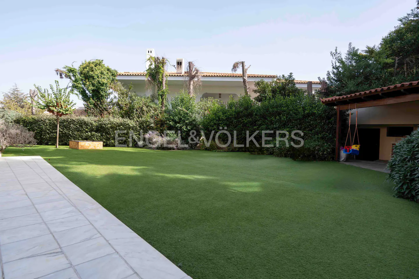 A lush green lawn with trees and bushes leads to a white house under a blue sky. An Engel & Völkers logo is superimposed on the lawn.