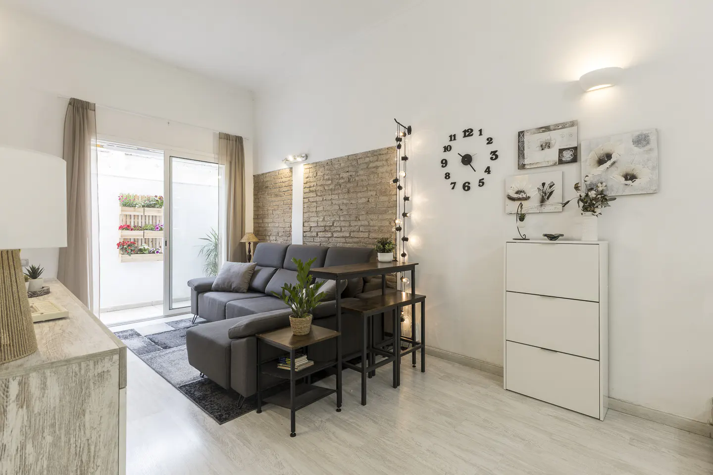 Bright living room with a gray sectional sofa, brick accent wall, and white storage cabinet. A sliding glass door leads to a patio with flowers.