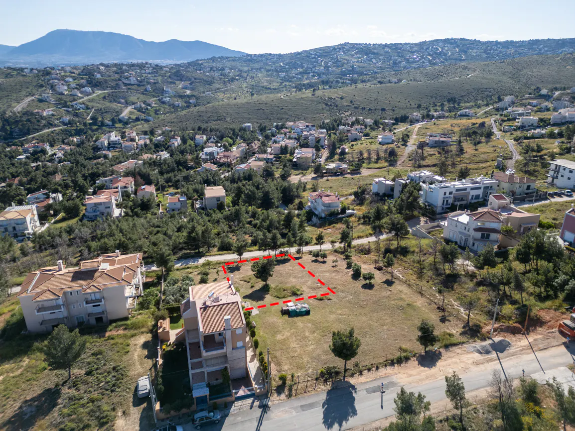 Aerial view of a vacant lot outlined in red, surrounded by houses and trees, with mountains in the background.