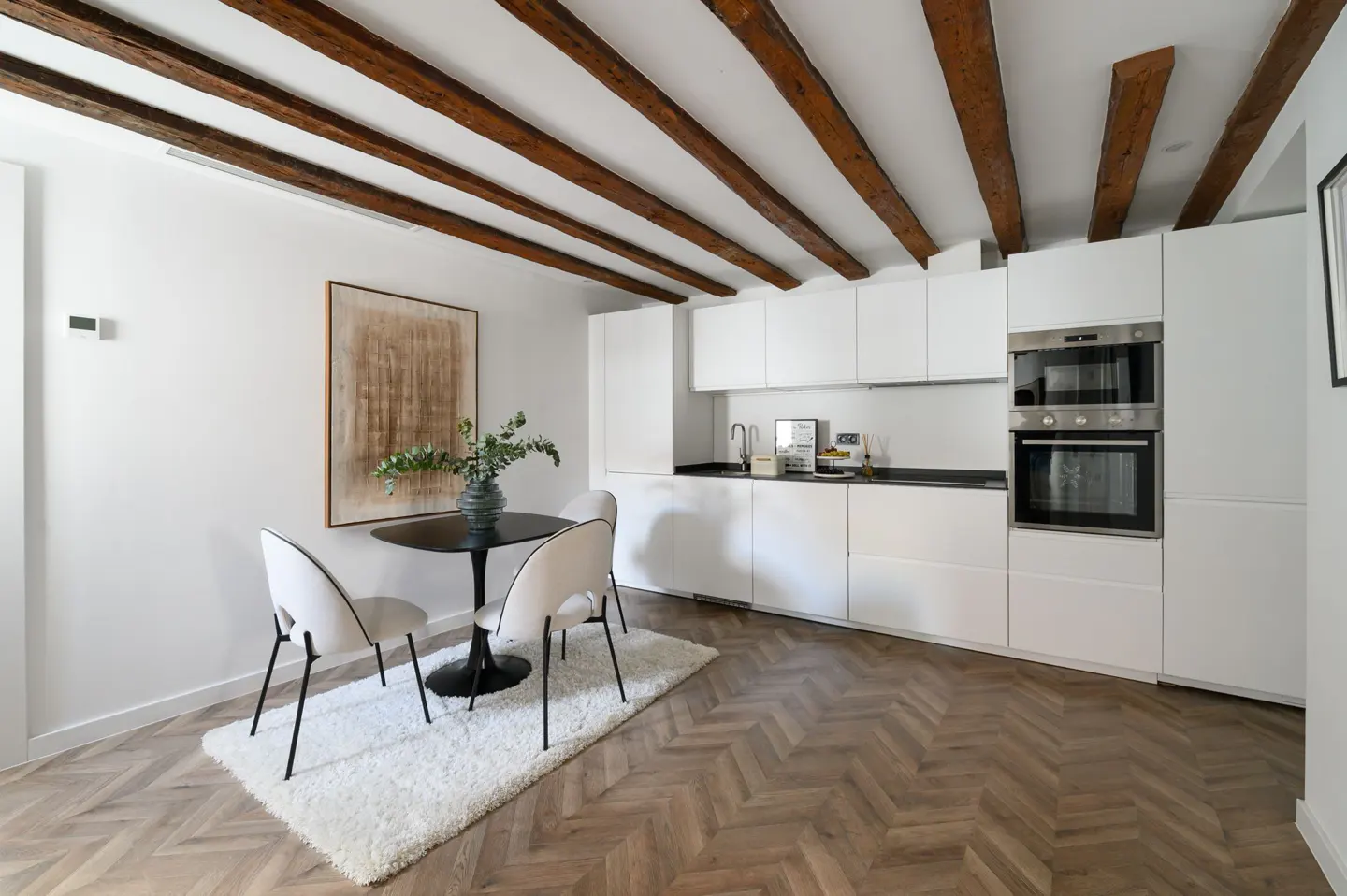 Bright kitchen with white cabinets, black countertops, and wood beams. A round table with chairs sits on a white rug.