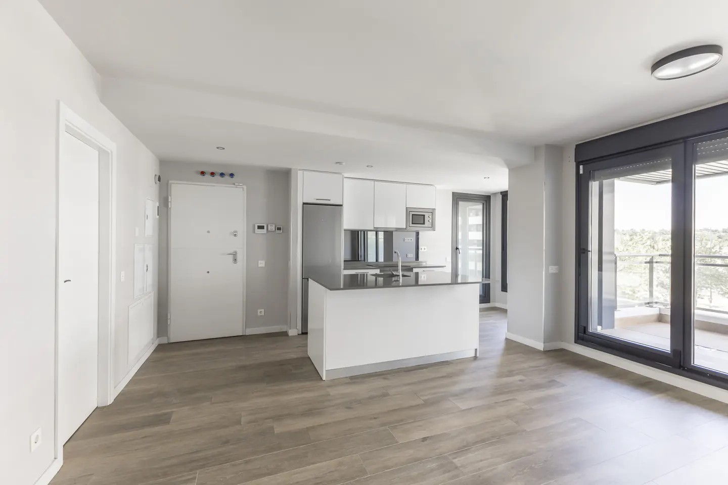 Bright, modern apartment with wood floors, white walls, and an open kitchen with a dark countertop island. Sliding glass doors lead to a balcony.