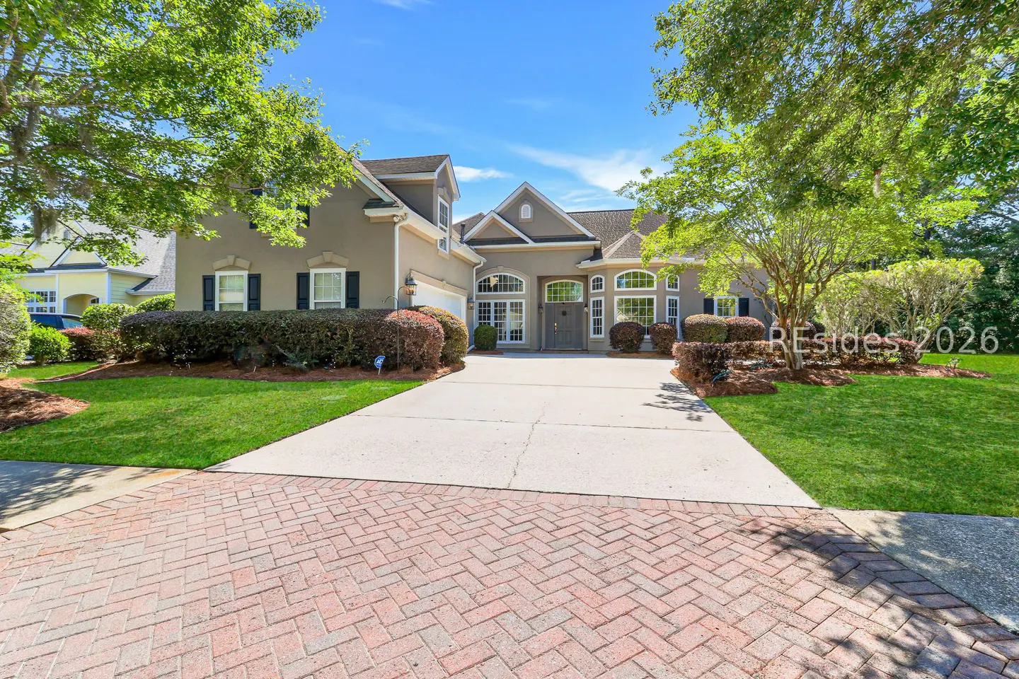 A two-story tan house with a gray roof, black shutters, and a brick and concrete driveway.