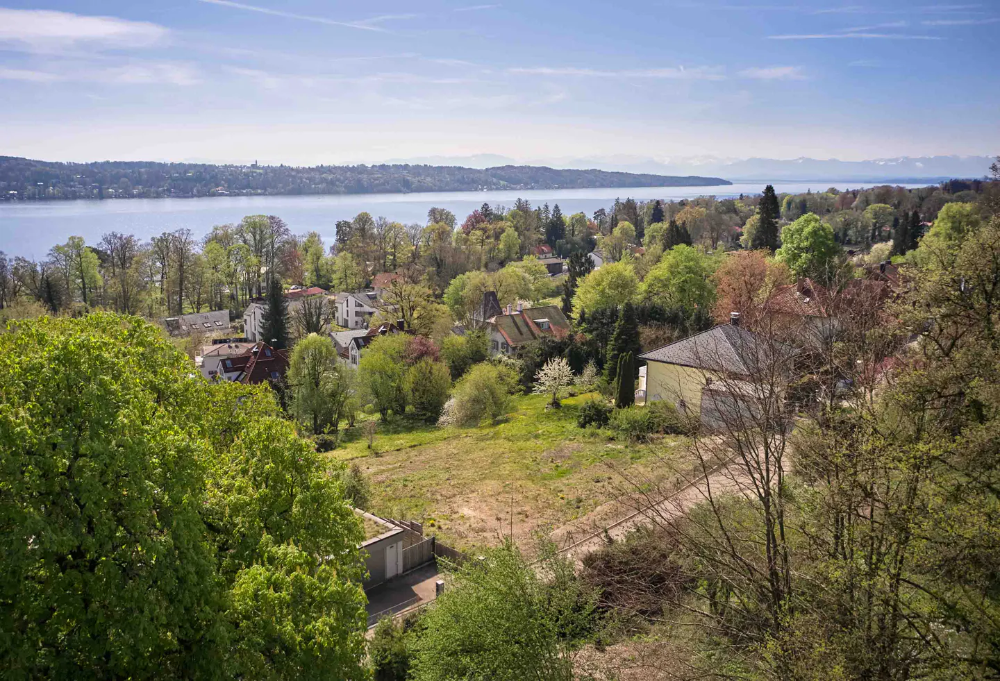 Scenic view of houses nestled among green trees, overlooking a blue lake and distant mountains under a bright sky.
