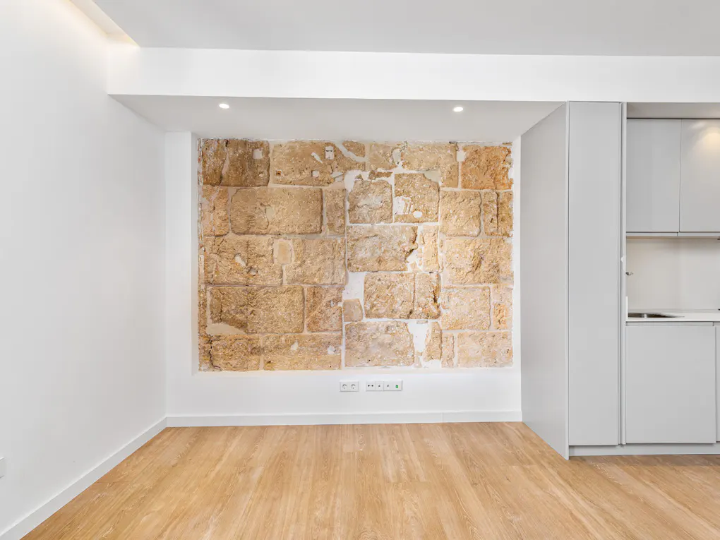 Minimalist apartment interior with a tan stone accent wall, light wood floors, and white walls. A modern kitchen is visible on the right.