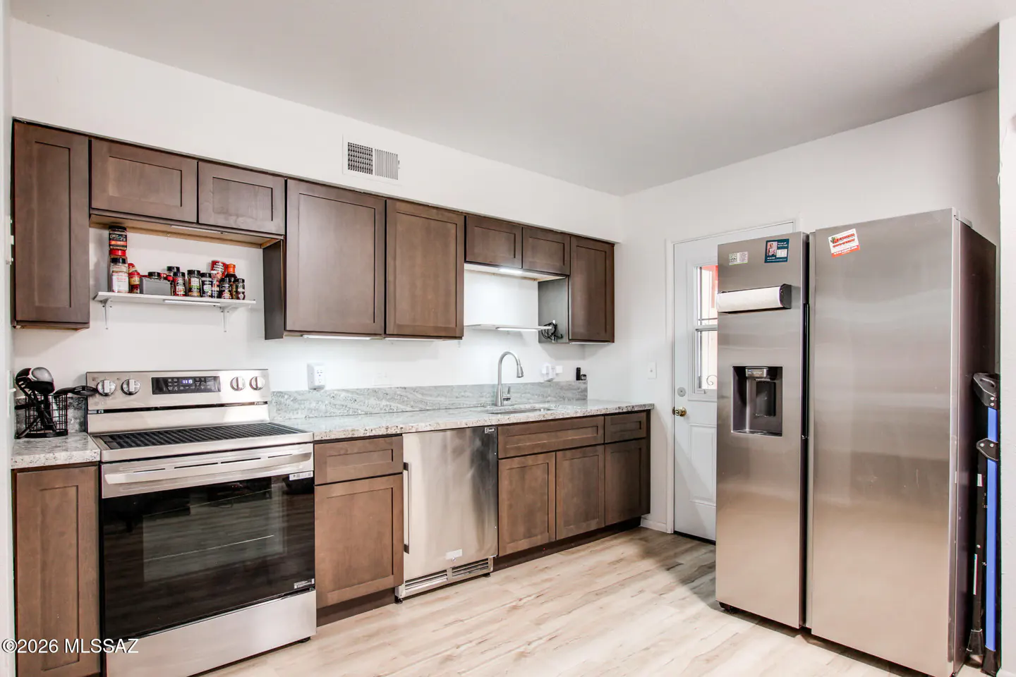 A kitchen with brown cabinets, stainless steel appliances, and light wood-look flooring. A white door is visible in the background.