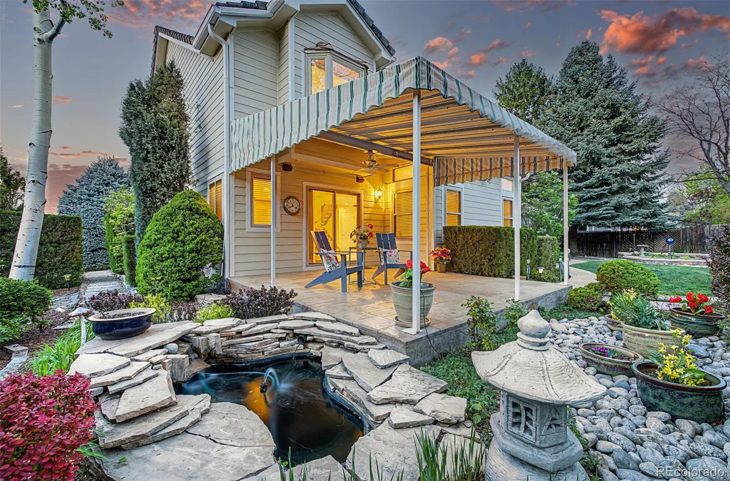 Backyard view of a beige house with a striped awning, patio furniture, and a rock-lined pond with a small fountain.