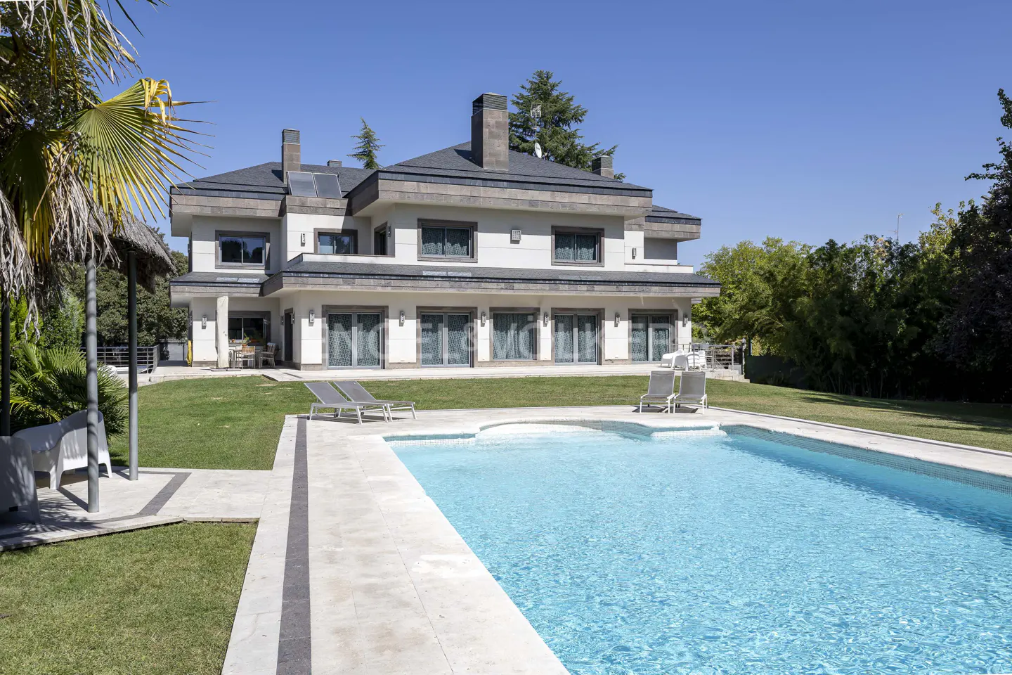Large two-story luxury home with a swimming pool, lounge chairs, and a green lawn under a clear blue sky.