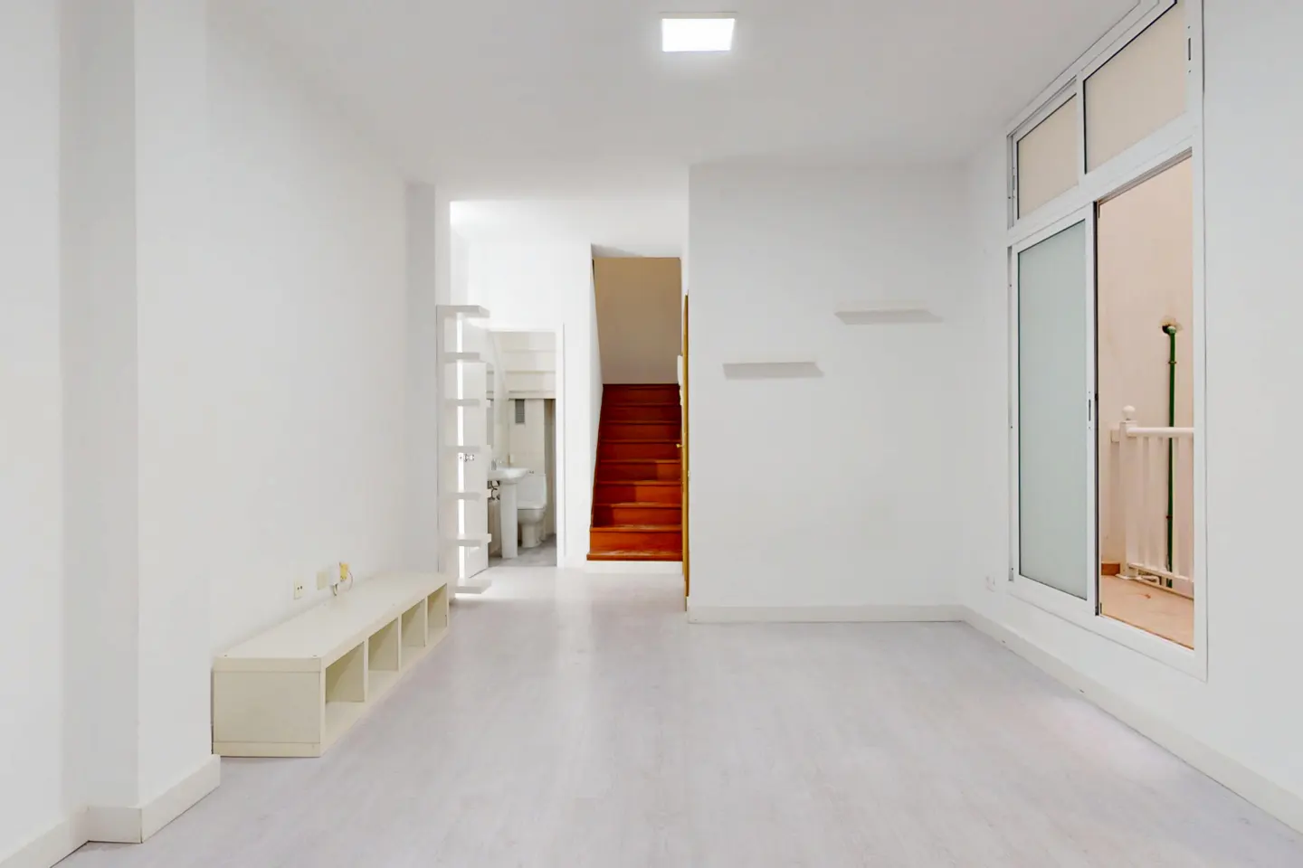 Bright, empty room with white walls, light wood floors, and a white storage unit. A staircase and bathroom are visible in the background. A sliding glass door is on the right.