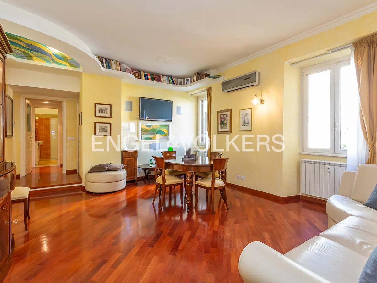 Bright living room with hardwood floors, yellow walls, and a round wooden table with chairs. A white sofa sits near a window with beige curtains.