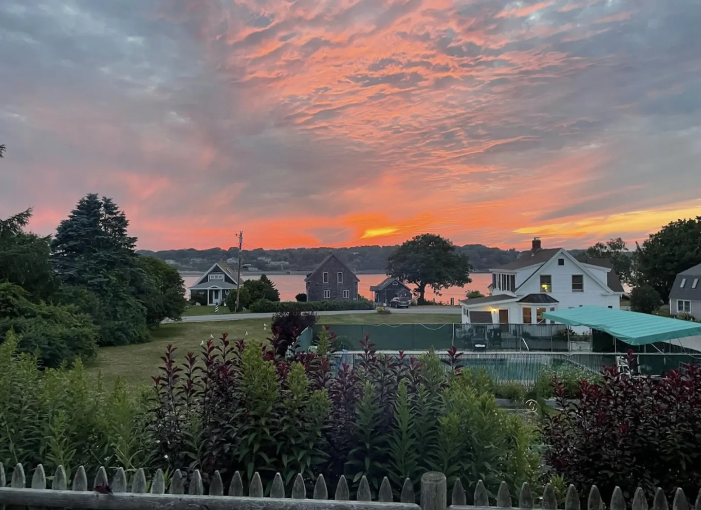 Sunset view over water with houses, trees, and a pool. The sky is pink and orange. A white fence and plants are in the foreground.