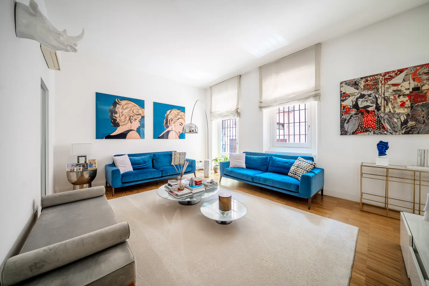 Modern living room with two blue sofas, a grey daybed, glass coffee tables, and vibrant pop art on white walls.