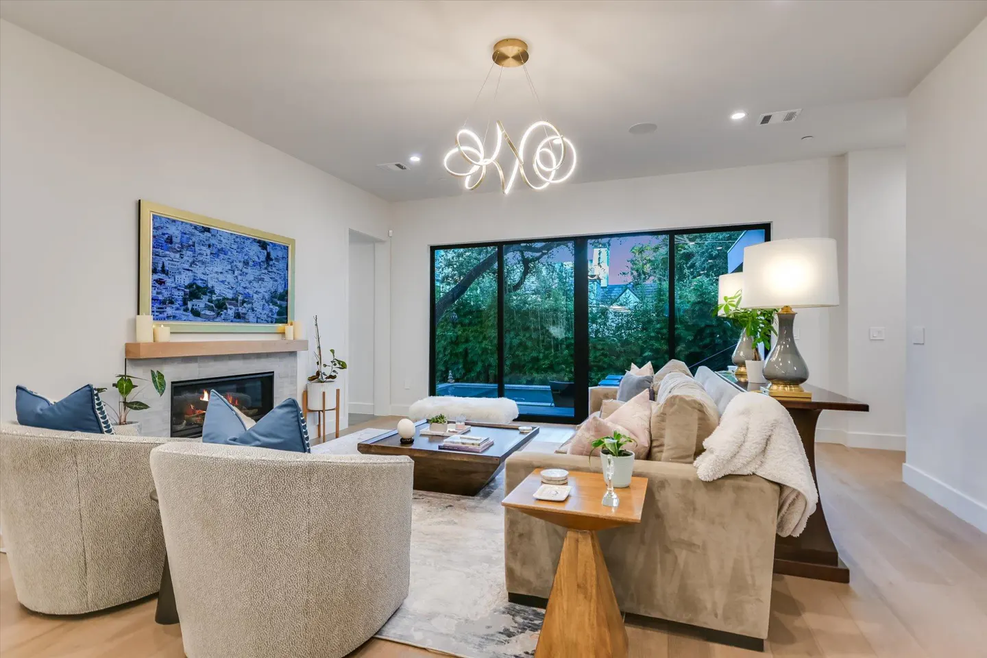 Bright living room with beige chairs, sofa, and a fireplace. A modern chandelier hangs above, and large windows offer a view of greenery.