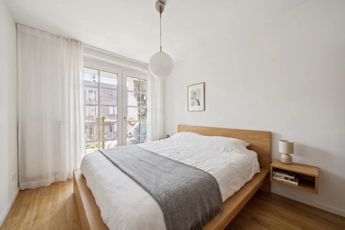 Bright bedroom with a wooden platform bed, white linens, and a gray throw blanket. French doors lead to a balcony.