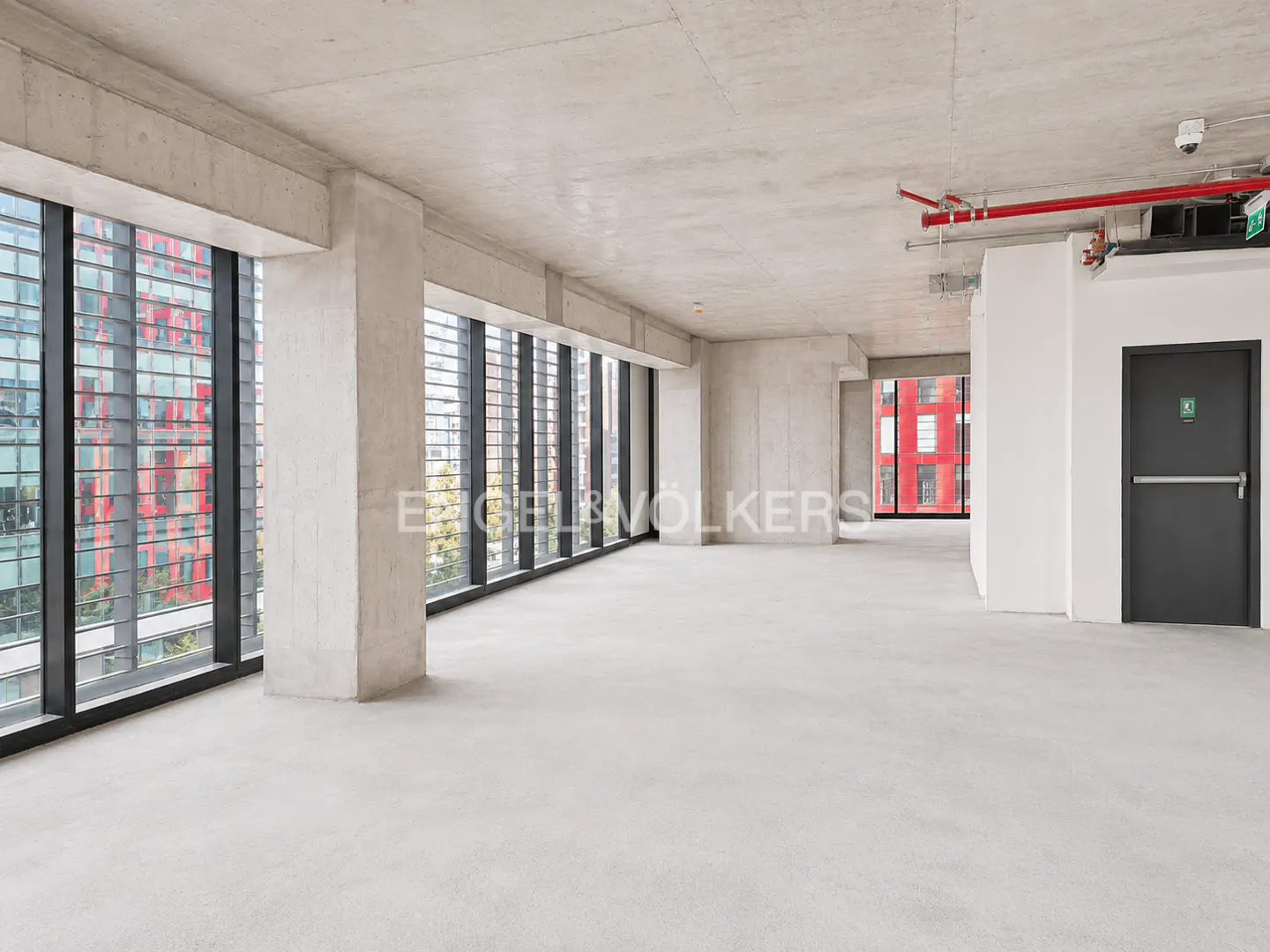 Unfurnished office space with concrete floors, pillars, and ceiling. Large windows with black frames offer city views. A black door is on the right.