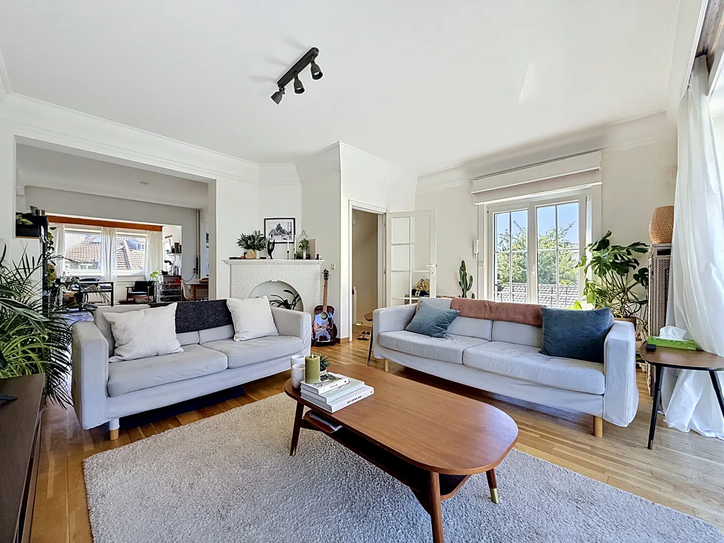 Bright living room with two light gray sofas, a wood coffee table on a gray rug, and hardwood floors. A guitar leans against a white fireplace.