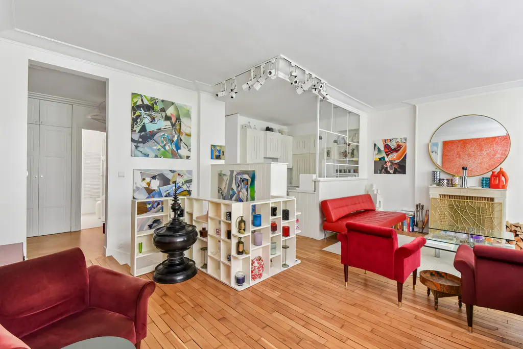 Bright living room with hardwood floors, red velvet chairs, and white shelving. Abstract art adorns the white walls. Kitchen visible in the background.