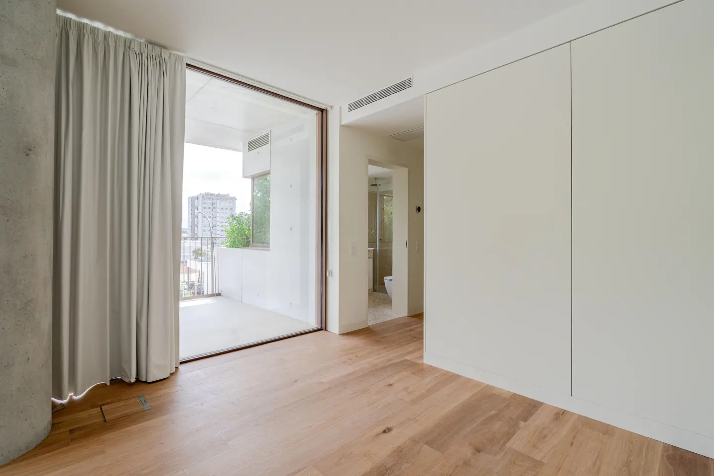 Bright room with wood floors, white walls, and a balcony. Gray curtains are partially drawn. A bathroom is visible through a doorway.