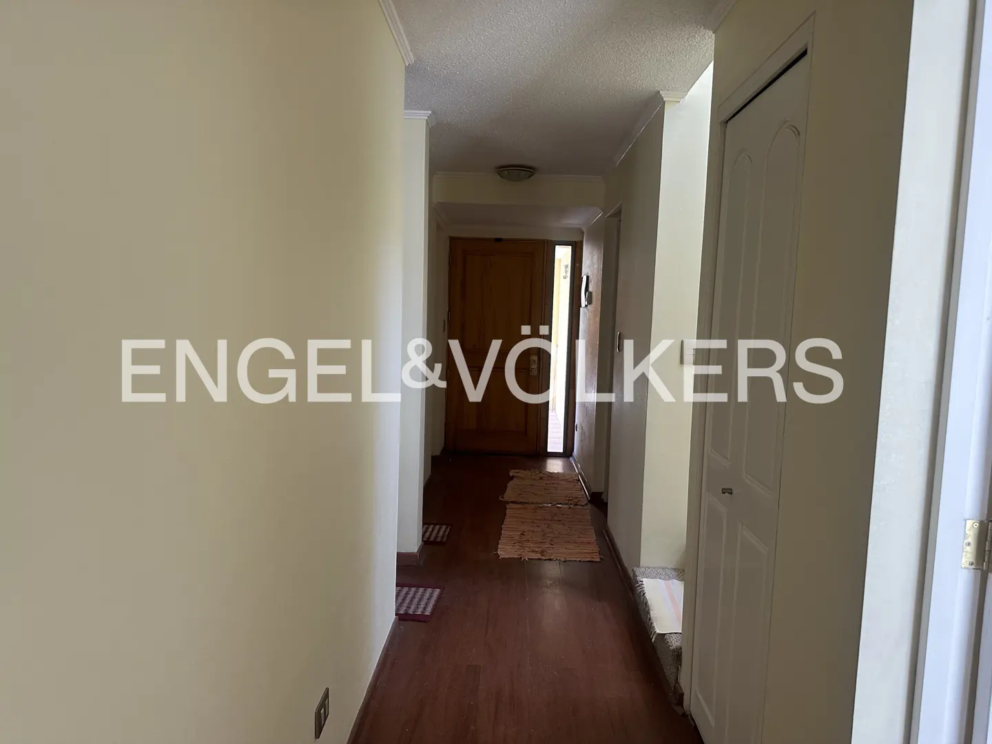 Hallway with wood floors, beige walls, and white trim. A brown door is at the end of the hall. Engel & Völkers logo is visible.