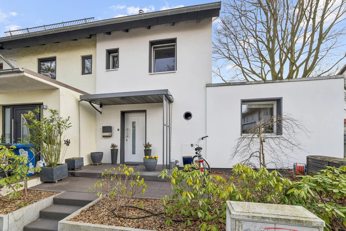 Modern white townhouse with a gray awning over the front door, potted plants, and a bicycle parked nearby.