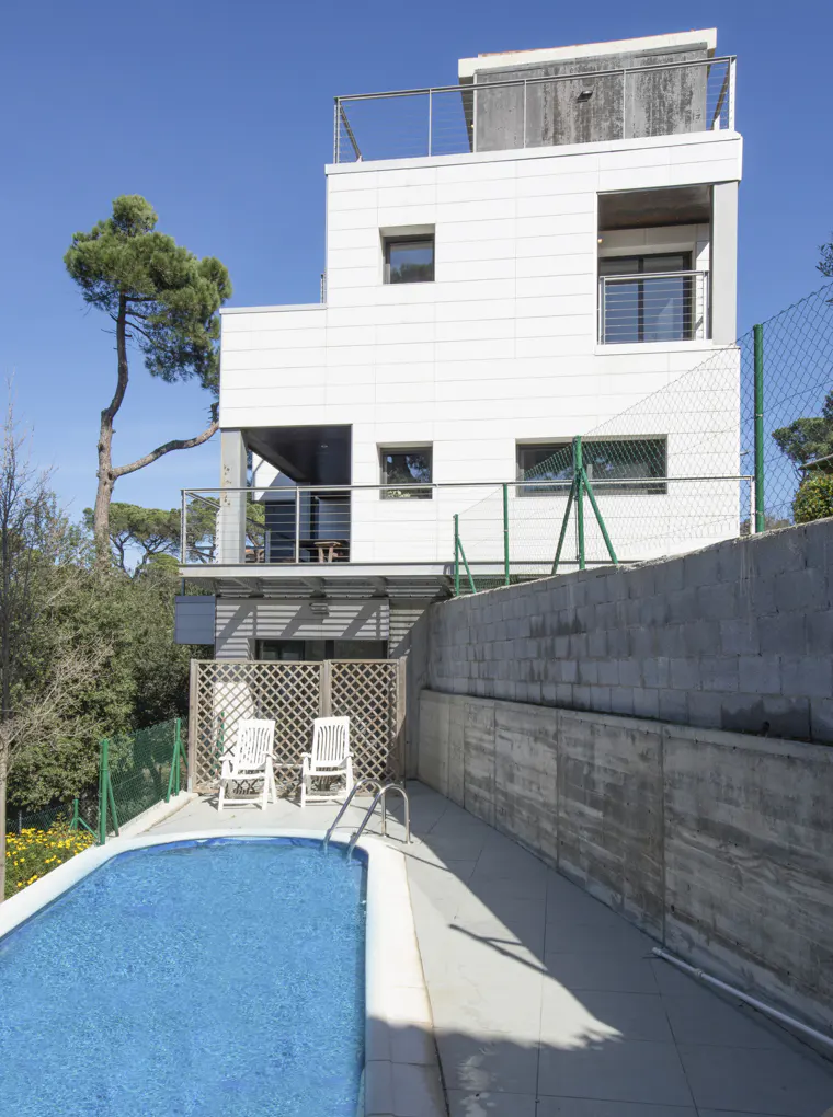 Modern white house with a blue pool, two white chairs, and a green fence under a clear blue sky.