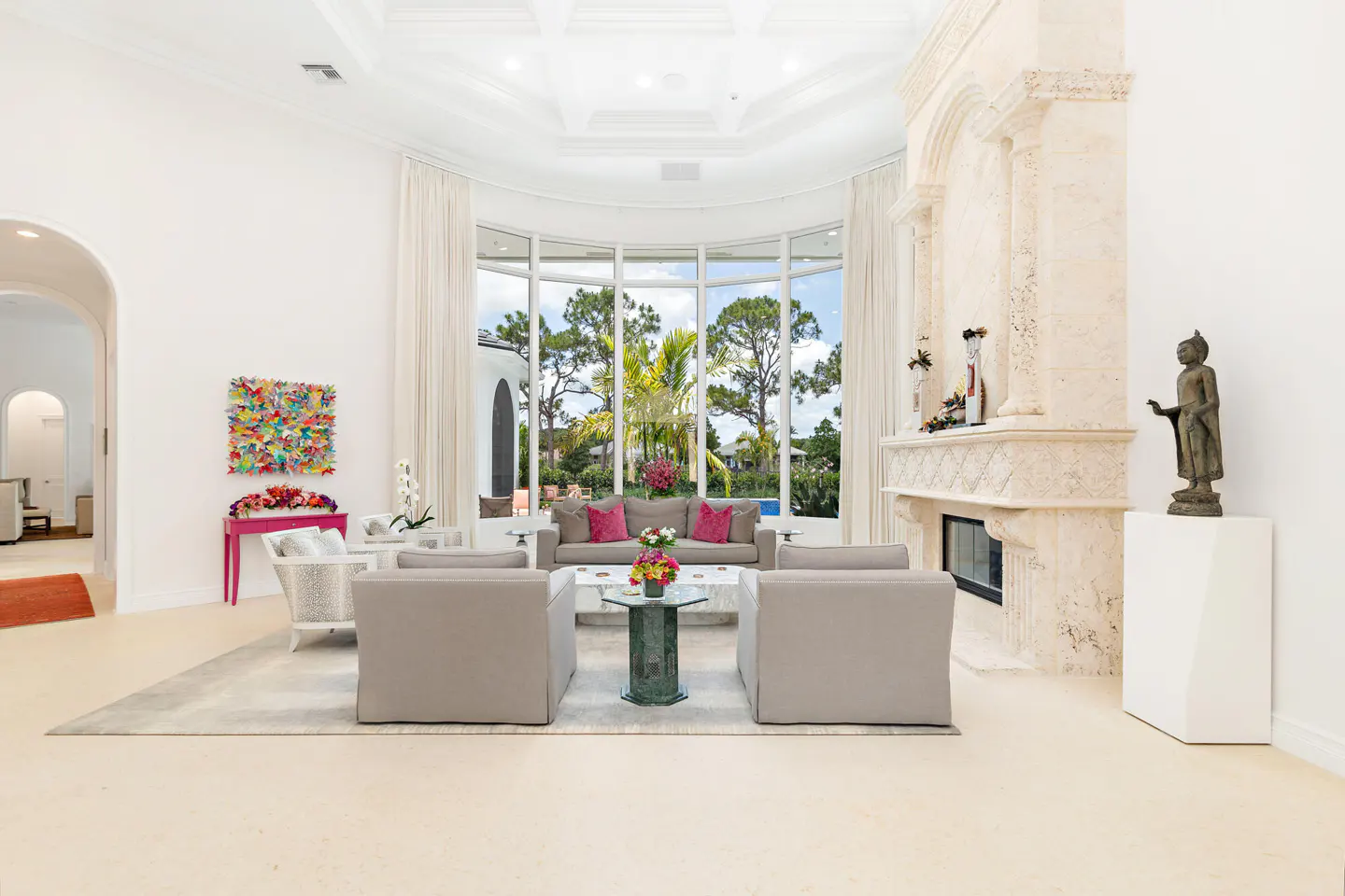 Bright, airy living room with beige chairs, sofa, and a stone fireplace. Large windows overlook lush greenery. A Buddha statue stands near the fireplace.