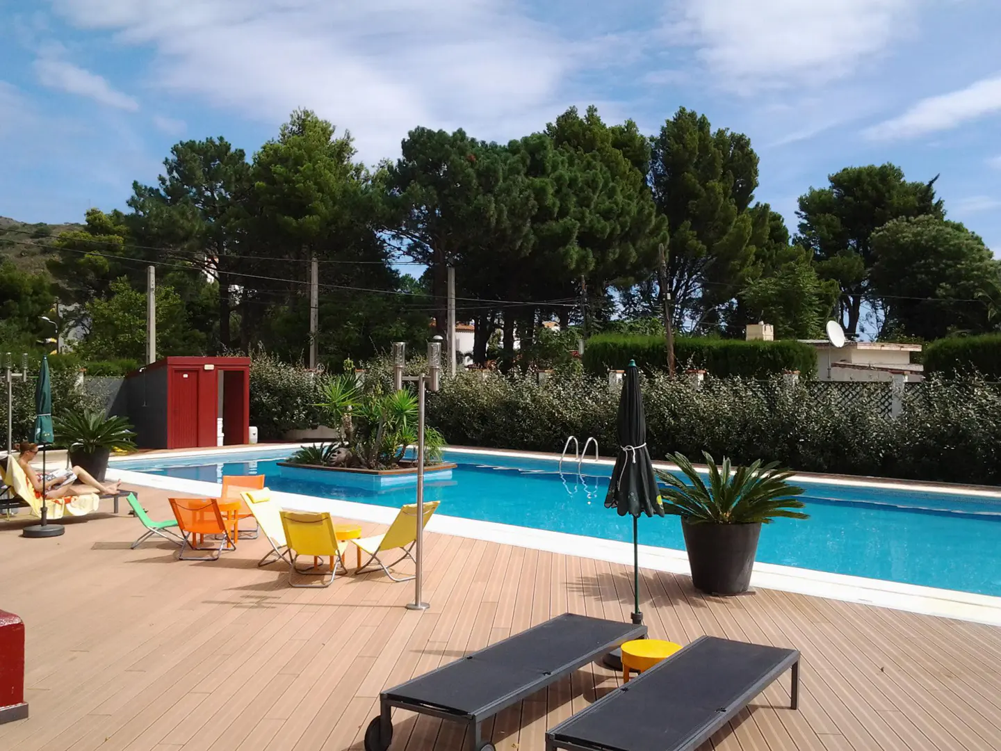 A sunny outdoor pool area with lounge chairs, colorful chairs, and a woman reading on a lounger. Trees and a blue sky are in the background.