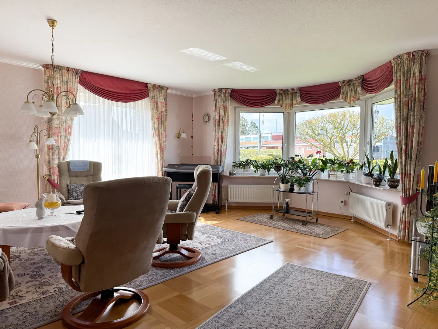 A bright living room with hardwood floors, floral curtains, and two beige armchairs. A piano and plants are near the window.