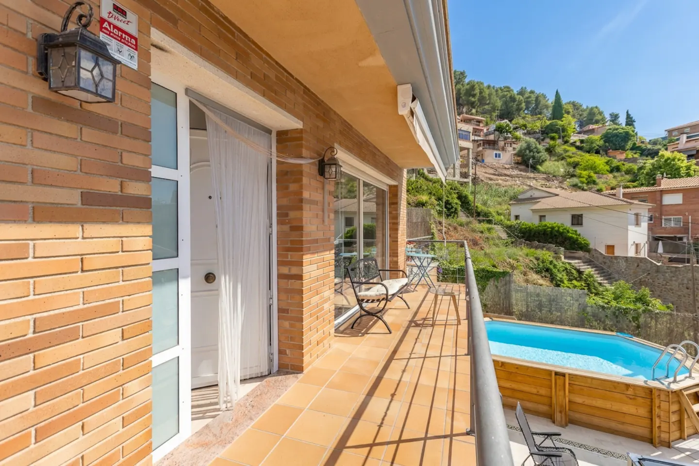 Balcony view of a brick house with a pool. Chairs and a table sit on the tiled balcony, overlooking a blue pool and a green hillside.