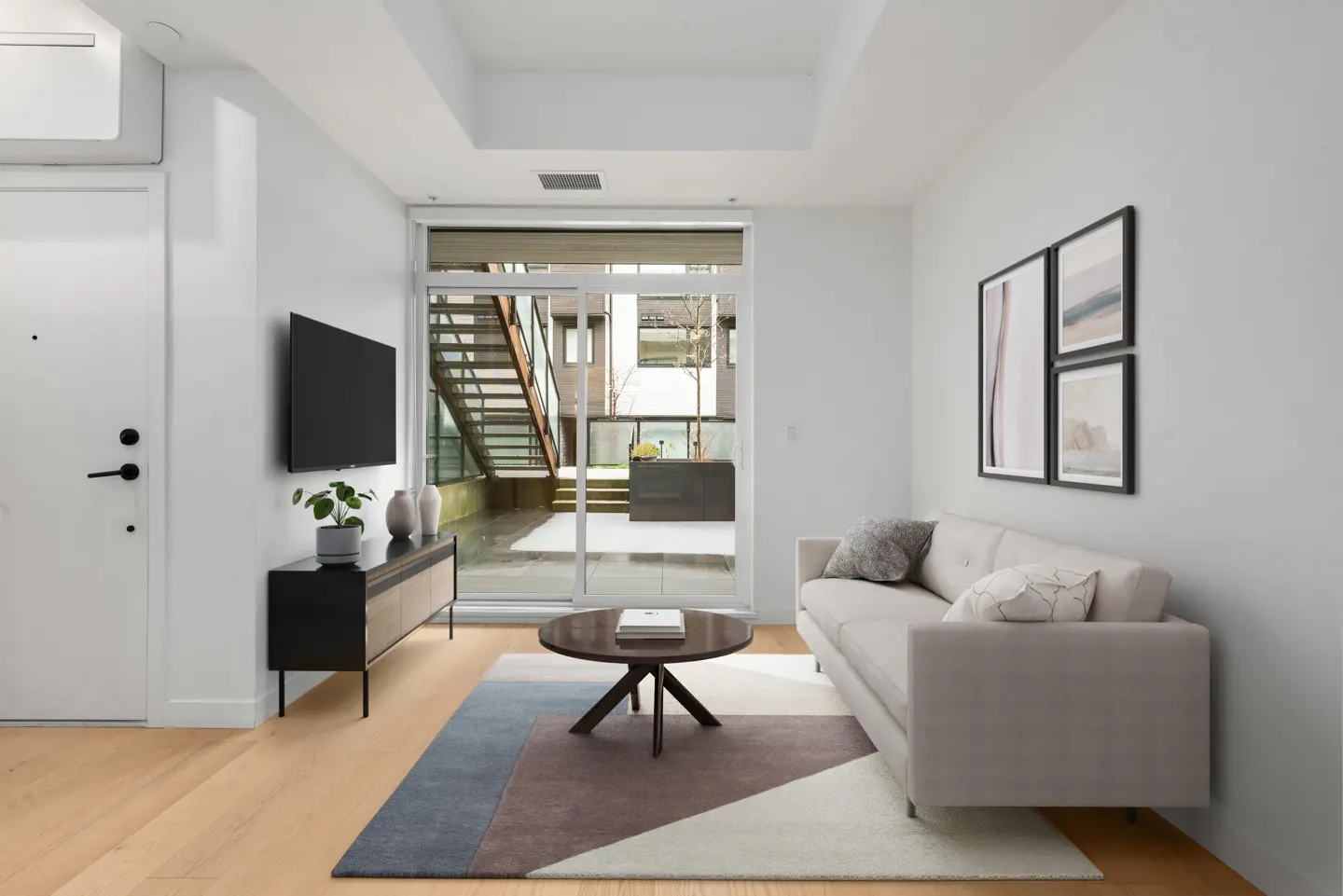 Bright living room with wood floors, a gray sofa, and a round coffee table on a geometric rug. A sliding glass door leads to an outdoor staircase.