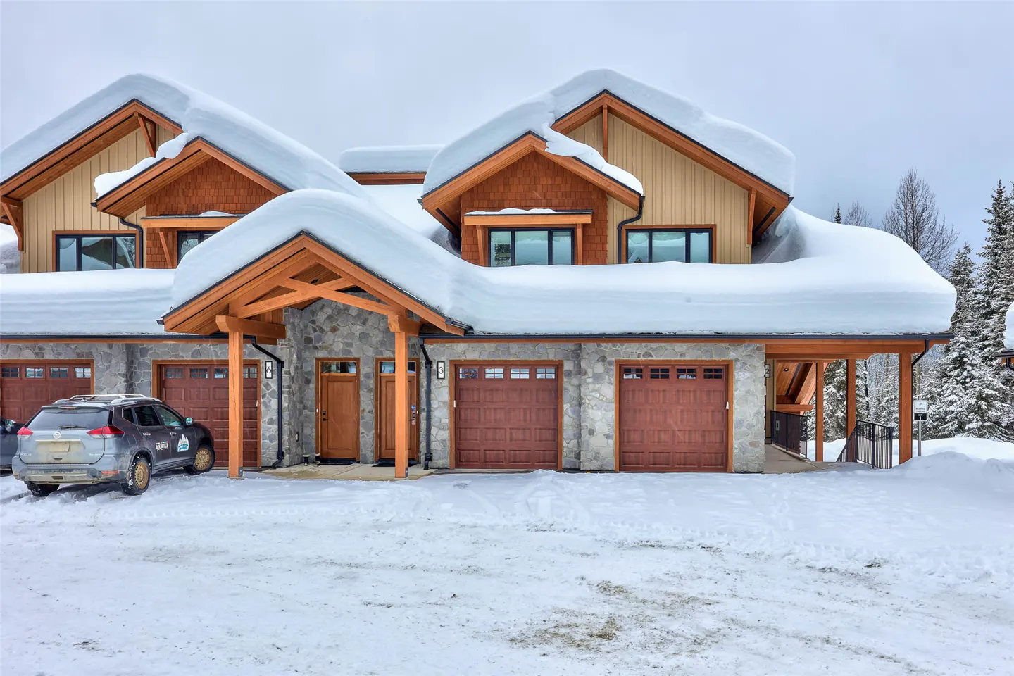 A snow-covered townhouse with stone and wood accents, brown garage doors, and a car parked in the snowy driveway.