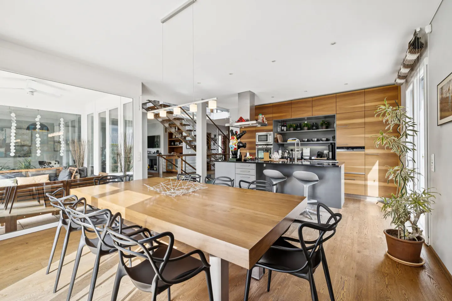 Open-concept dining and kitchen area with a large wooden table, black chairs, and modern kitchen cabinets. A staircase is visible in the background.