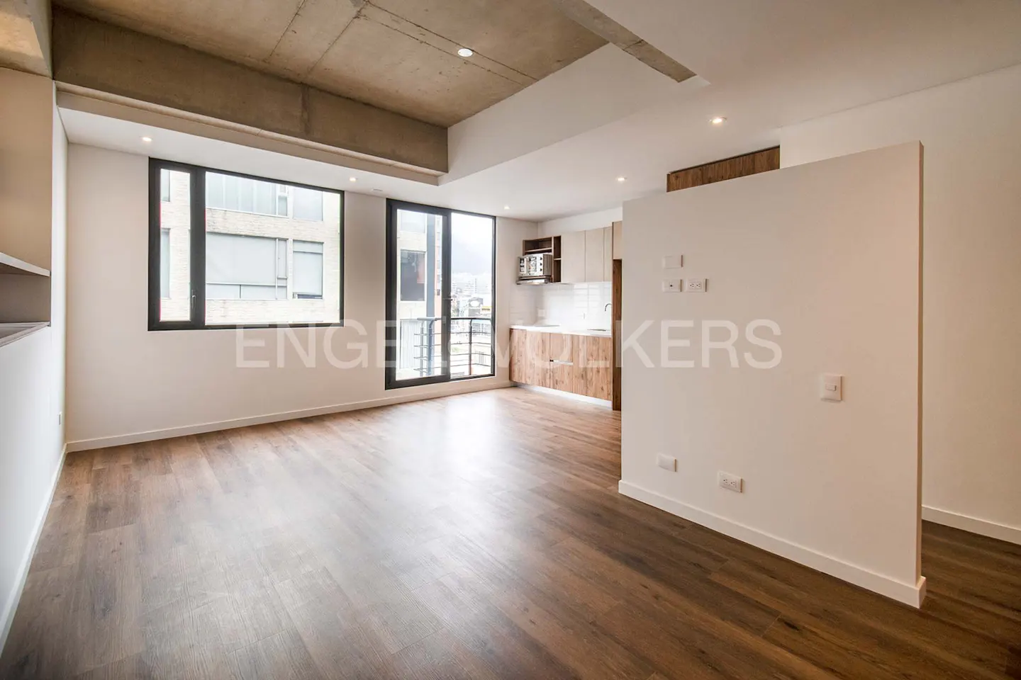Bright, modern apartment interior with wood floors, white walls, and a concrete ceiling. Large windows offer city views. A kitchen is visible in the background.