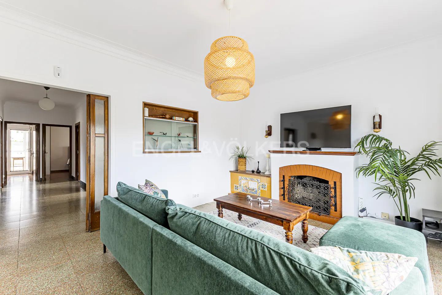Living room with green sofa, wood table, and fireplace with TV above. A woven light fixture hangs from the ceiling. Hallway in background.