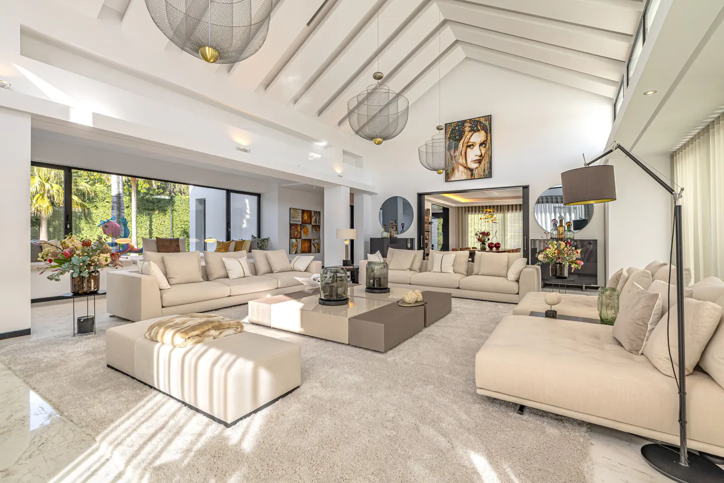 Bright, open living room with vaulted ceilings, beige sofas, and a large gray coffee table. Modern light fixtures hang from the ceiling. A portrait hangs on the wall.