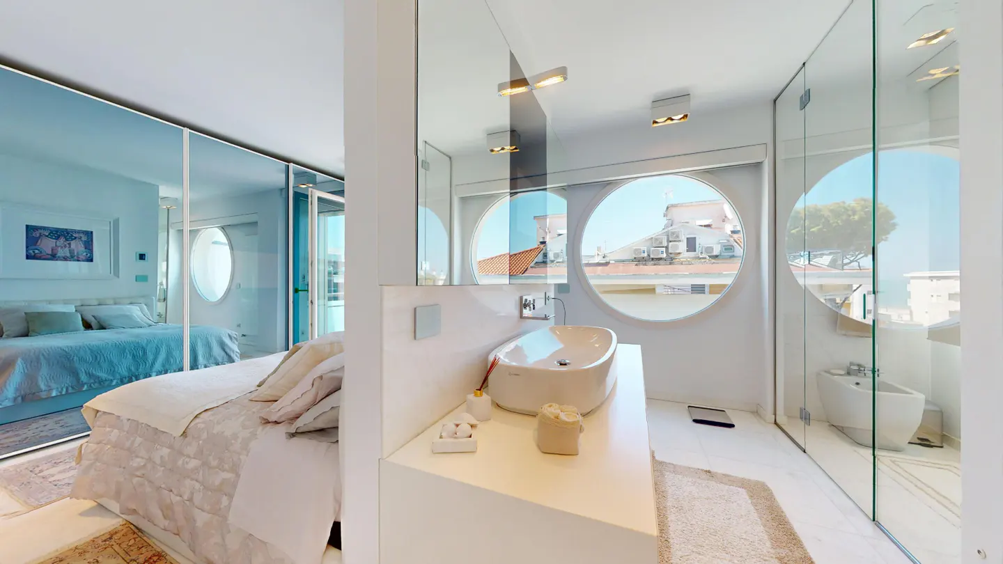 Bright, modern bathroom with a white sink, round windows, and glass shower. A bedroom is visible through a glass wall.