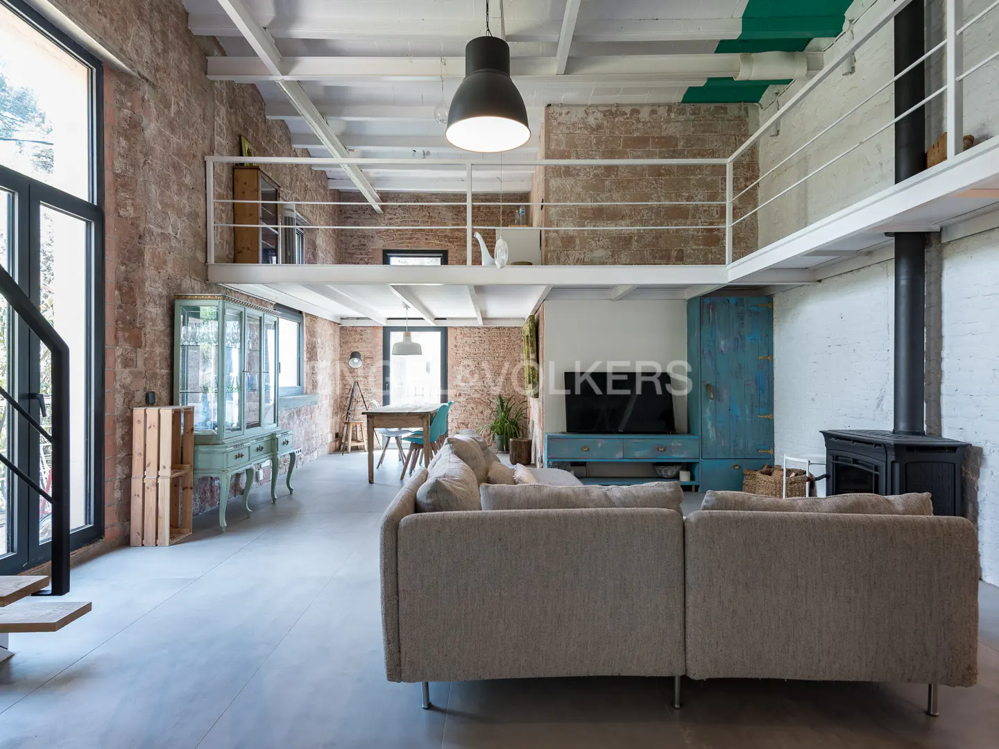 A modern living room with exposed brick walls, a gray sofa, and a black fireplace. A loft with white railings overlooks the space.