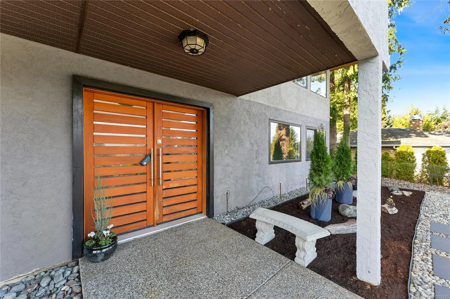 Exterior view of a modern home with a wood slat double door, gray stucco, and a stone bench.