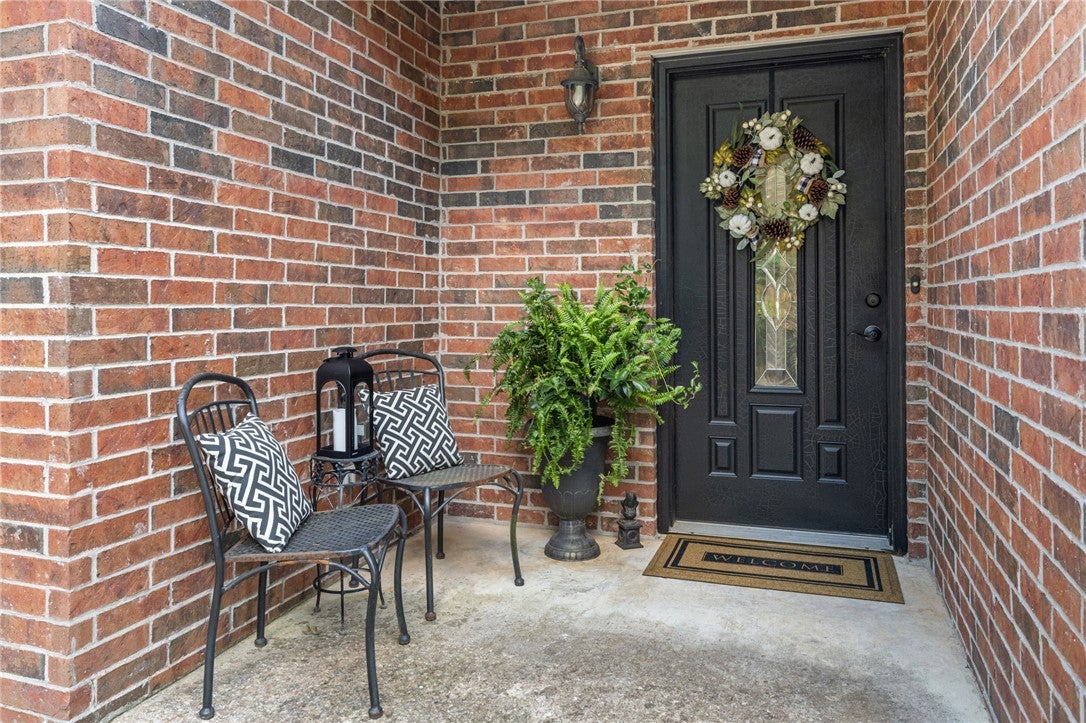 Black front door with wreath, two chairs with patterned pillows, a potted fern, and a welcome mat on a brick porch. [AI-generated text]