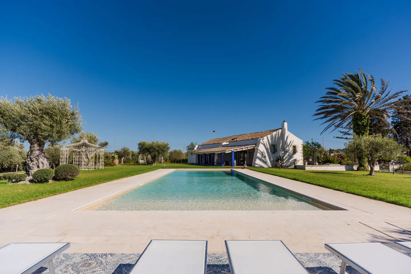 A sunny view of a white house with a pool, lawn, and trees. Lounge chairs are in the foreground.