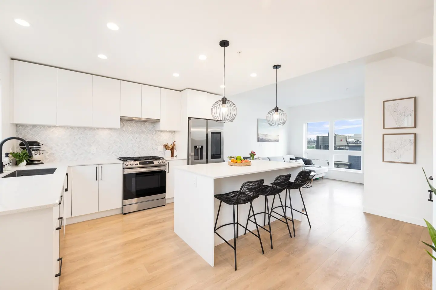 Bright, modern kitchen with white cabinets, stainless steel appliances, and a large island with black bar stools. Living room visible in the background.