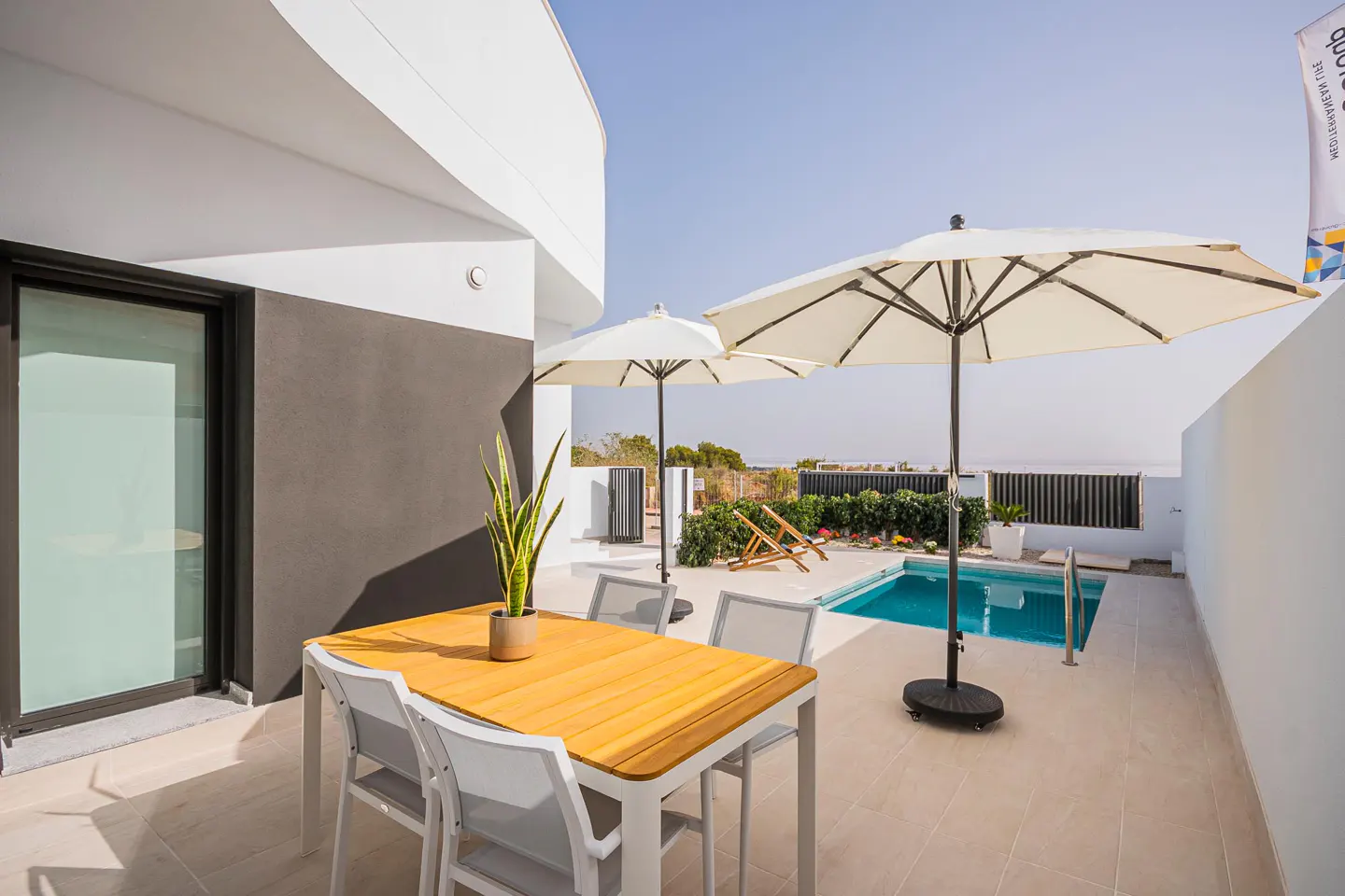 Outdoor patio with a wooden table, white chairs, umbrellas, and a pool. A potted plant sits on the table.