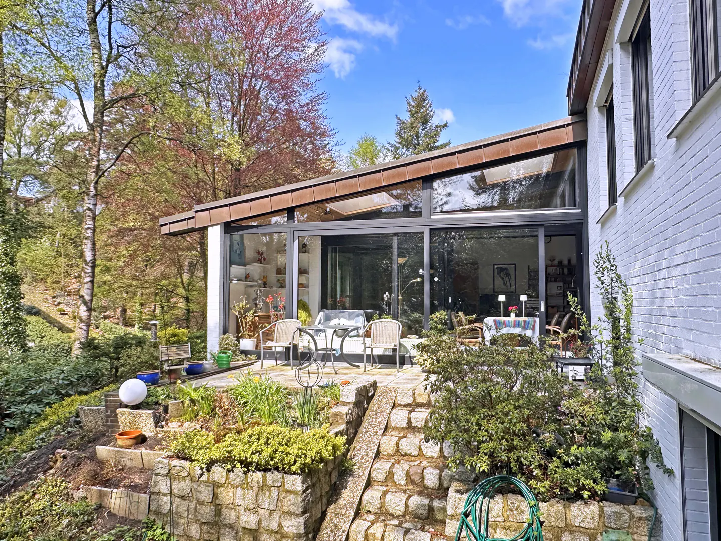 Exterior view of a modern white brick home with a glass-enclosed sunroom and stone patio surrounded by lush greenery and trees.