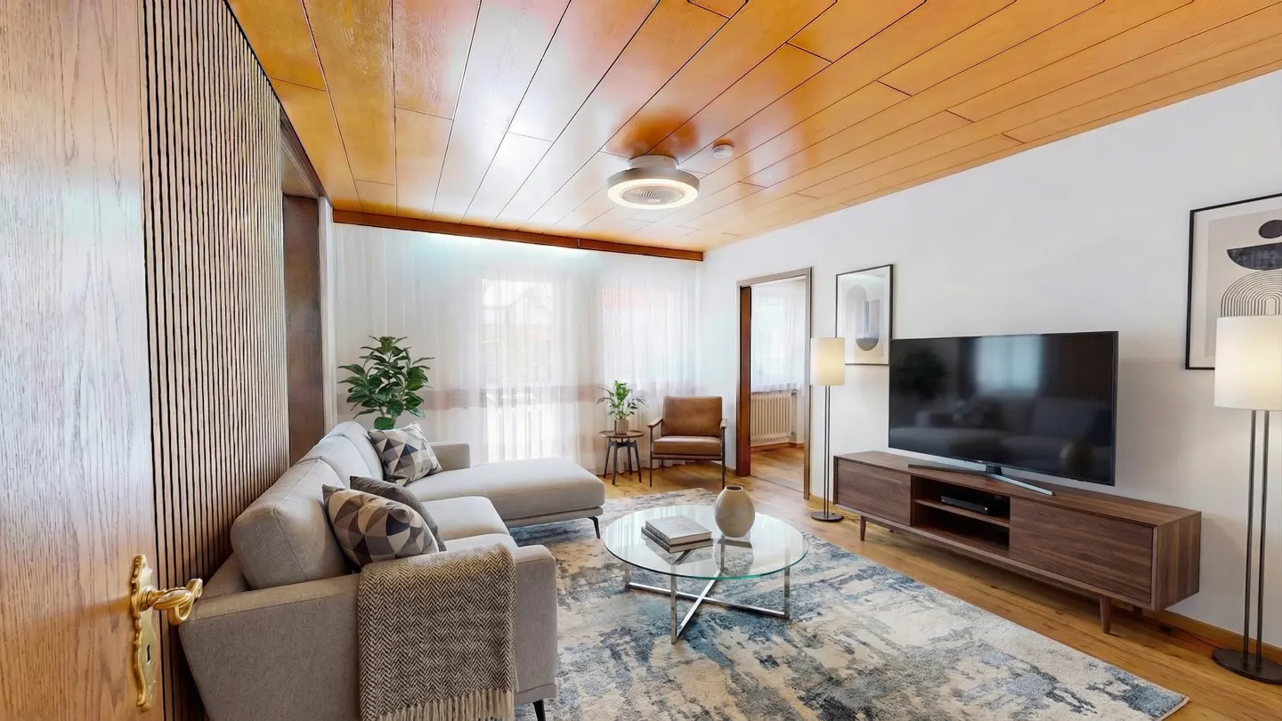 A modern living room with a gray sectional sofa, glass coffee table, and wood-paneled ceiling. A large TV sits on a wooden console.