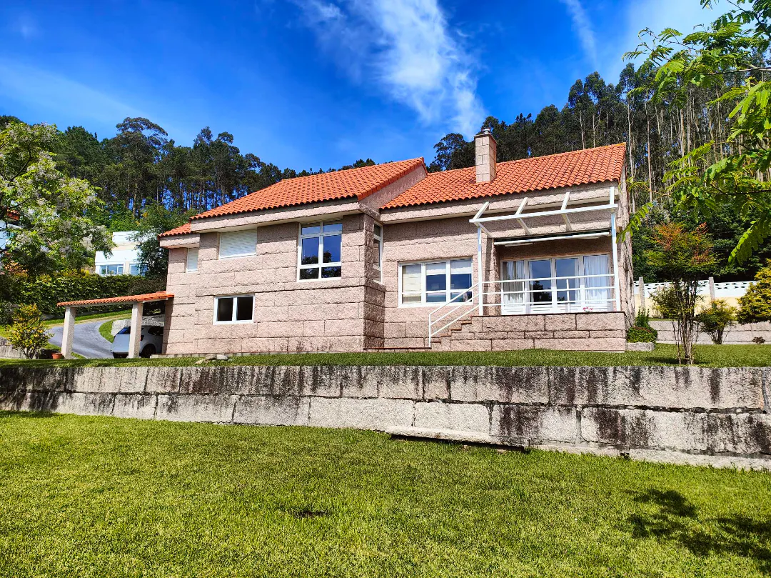 A two-story stone house with a red tile roof sits on a green lawn under a blue sky. Trees are in the background.