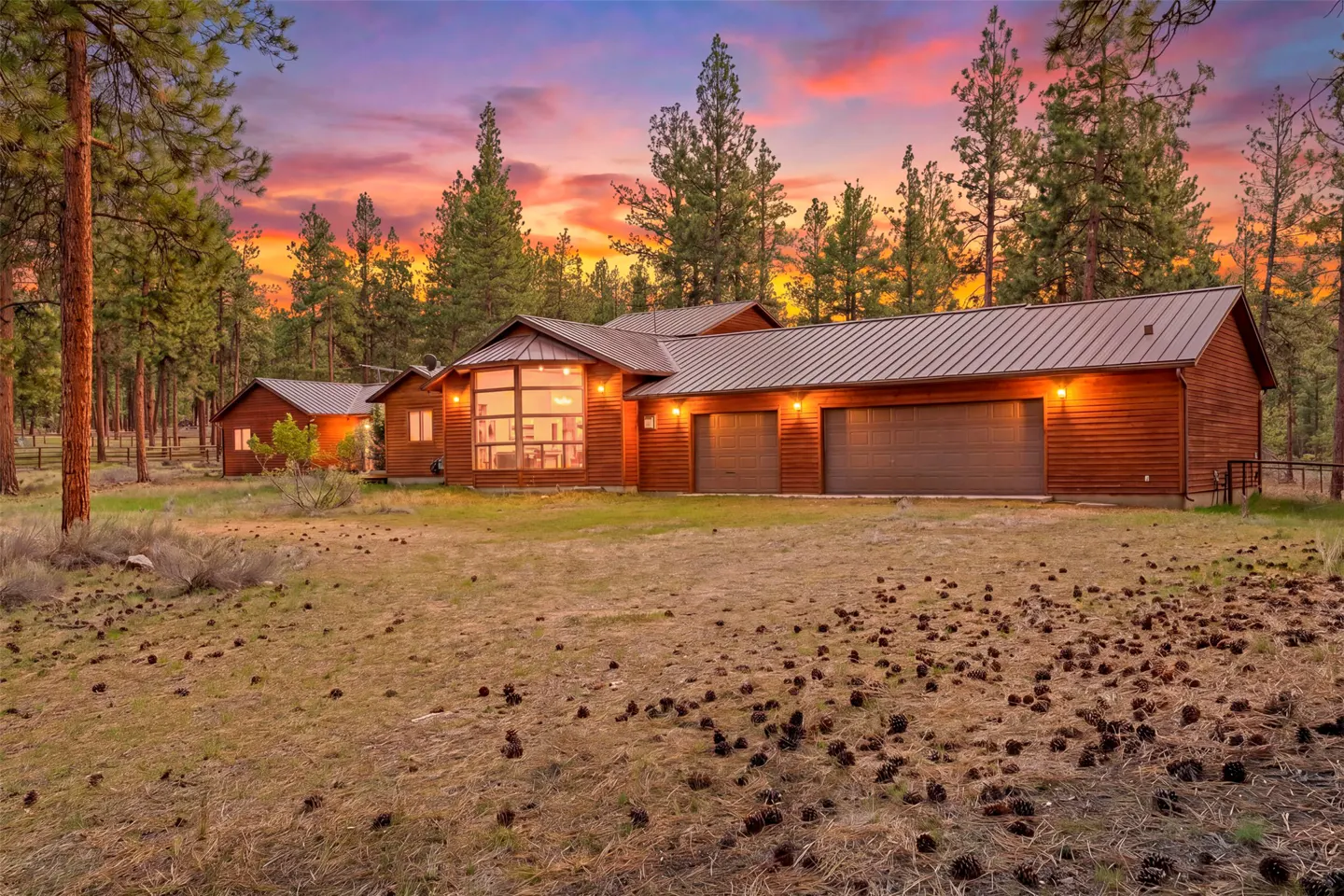 A log cabin-style house with a metal roof is surrounded by tall pine trees and a sunset sky.