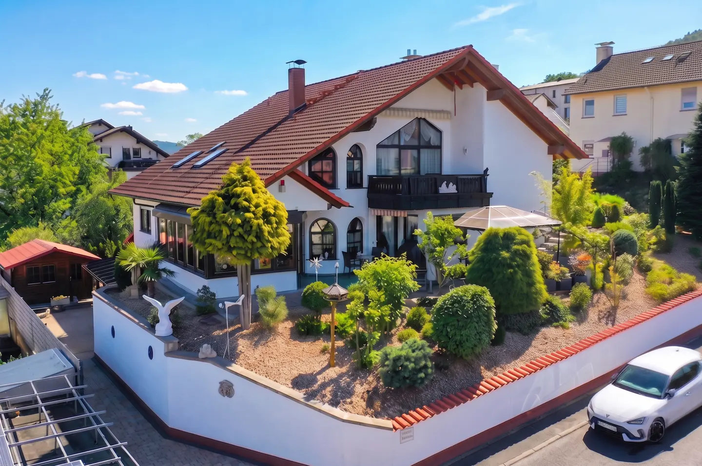 Exterior view of a white two-story house with a red tile roof, balcony, and landscaped garden with a white car parked nearby.